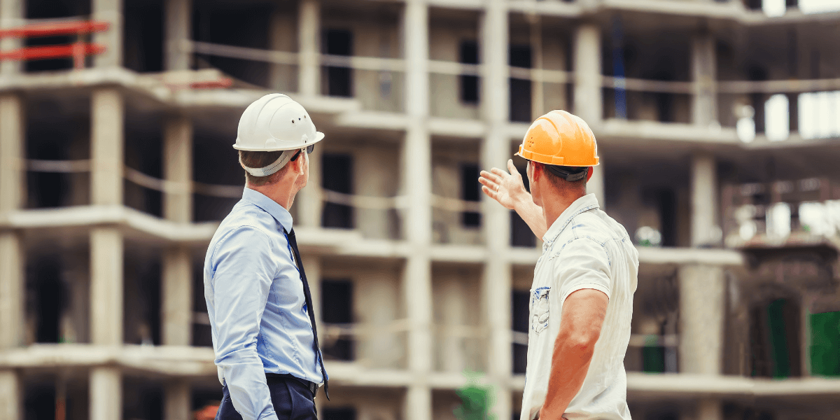 Two construction professionals wearing hard hats discussing plans in front of a multi-story concrete building under construction.